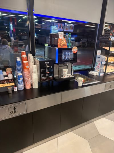 Here is a detailed description of the image:
The image appears to be taken inside a cafe or restaurant, likely near a serving counter. Various items are visible on the counter, including stacks of paper cups in different colors (white, orange, and blue), packets of sugar, stir sticks, and what appears to be a coffee machine. Behind the counter, there's a glass display case with reflections of the outside street, including building lights and signs. The reflections make it difficult to discern specific details about the exterior. The image is taken from a high angle, looking down onto the counter. The location appears to be in Stara Pazova Municipality, Serbia, based on information from the menu displayed on the coffee machine.