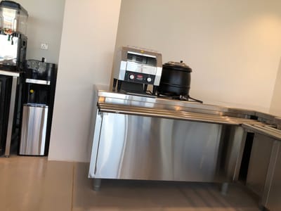 The image displays an interior view of a commercial food service area, located in the city of Hanak, Saudi Arabia. On the left side of the frame, a black and stainless steel beverage dispenser, featuring a clear top reservoir containing what appears to be sliced fruit, is situated next to a black and silver water cooler/dispenser with a lower stainless steel bin. A white electrical outlet is visible on the light-colored wall behind these appliances.
To the right, a multi-section stainless steel serving counter or buffet line extends horizontally. Positioned on this counter are a stainless steel commercial toaster, likely a conveyor model, equipped with a black control panel and red indicator lights, and a black, lidded soup tureen resting on a stand. The walls are uniformly light-colored, and the floor is a smooth, light tan or beige surface. No individuals are present, and no actions or interactions are depicted. The scene is well-lit and organized, consistent with a clean, functional commercial kitchen or self-service dining environment.
