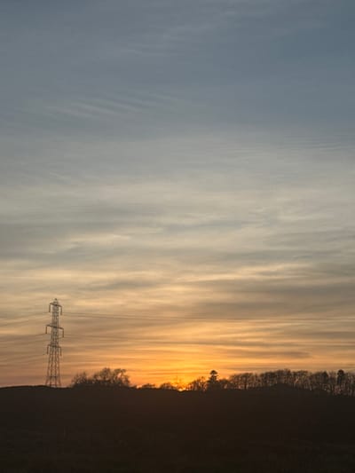 The image displays a rural landscape near Liff, United Kingdom, during either sunrise or sunset. The sky occupies the upper two-thirds of the frame, exhibiting a gradient from light grey-blue at the top to horizontal bands of orange and yellow near the horizon, consistent with a low sun angle. Cirrus or altocumulus clouds are visible as subtle, streaked patterns across the sky. In the left mid-ground, a multi-legged, metallic lattice-style electricity transmission pylon stands silhouetted against the illuminated sky, with multiple power lines extending horizontally to the right. The lower third of the image is dominated by a dark, undifferentiated landmass, representing ground or distant hills, topped by a dense, dark silhouette of a treeline along the horizon. The sun is partially visible as a bright orange glow directly above the treeline, slightly to the right of the frame's center. No individuals, specific actions, or interactions are discernible.