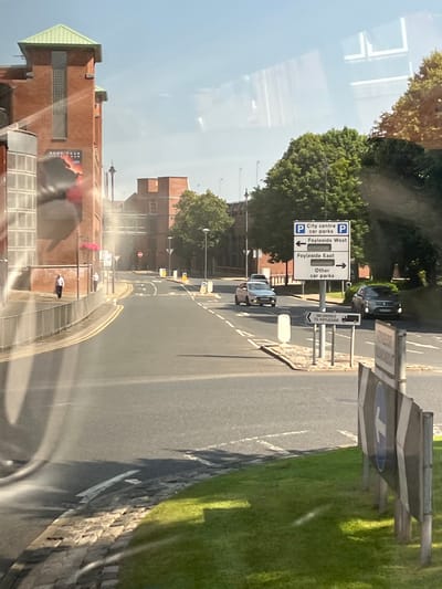 The image shows a street scene in the city of Derry/Londonderry, United Kingdom, on a sunny day. In the foreground, a gray car is visible on the right side of the road, facing forward. A speed limit sign with an arrow pointing left is also present. The road is paved and marked with white lines. On the left side of the road, a brick building with a green roof is visible, along with a sidewalk where two individuals are walking. 

A traffic signpost with multiple directional signs is located in the middle of the road. The signs indicate "City centre car parks," "Foyleide West" with an arrow pointing left, "Foyleide East" with an arrow pointing right, and "Other car parks" with an arrow pointing right. Another smaller sign below this indicates "DELIVERIES TO FOYLESIDE" with a truck symbol. Two more cars are visible in the distance, one silver and one gray. Trees line the side of the road, and the sky is clear and blue. The overall impression is that of a typical urban street environment.