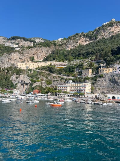 The image shows a scenic view of the coast in Pogerola, Italy. In the foreground, the clear blue water of the sea is calm with small ripples reflecting the sunlight. A number of boats, ranging from small motorboats to larger yachts, are anchored in a harbor. Some of the boats are white, while others are darker colored. Bright orange buoys are visible in the water.

Behind the harbor, a steep, rocky cliff face rises dramatically, covered in lush green vegetation and trees. Scattered along the cliffside are several buildings, including hotels and residences, with light-colored facades and red-tiled roofs. One prominent hotel building, with multiple stories and balconies, is situated directly above the harbor. Higher up the cliff, more buildings are visible, some appearing to be part of a larger complex or village. A winding road is carved into the mountainside, connecting different levels of the settlement. In the background, the clear blue sky suggests a bright, sunny day. The overall scene conveys a sense of a picturesque coastal town nestled into the dramatic Italian landscape.