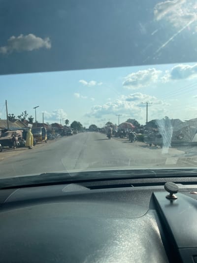 The image depicts a roadside scene in Oduape, Nigeria. The perspective is from inside a vehicle, looking out through the windshield. The road is lined with various objects and structures, suggesting a bustling market or residential area.

Prominent among the subjects are numerous motorcycles and a few tricycles, indicating their common use for transportation. These vehicles are parked along the side of the road and some are in motion further down. Small shops or stalls are visible, with some items displayed outside, hinting at commercial activity. There are also people present, though they are not clearly distinguishable due to the distance and resolution. Some structures appear to be simple buildings with roofs.

The scene is set in an outdoor environment under a bright sky filled with scattered white clouds. The lighting suggests it is daytime, likely mid-morning or early afternoon, with ample sunlight casting shadows. The weather appears to be fair.

Notable details include the presence of utility poles with wires stretching across the scene, a common sight in many populated areas. The windshield of the vehicle has some streaks and a small circular knob attached to the dashboard is partially visible in the foreground on the right side of the frame. The image quality is somewhat reduced by glare and the angle, making it challenging to discern fine details or specific emotions. There is no visible text in the image.