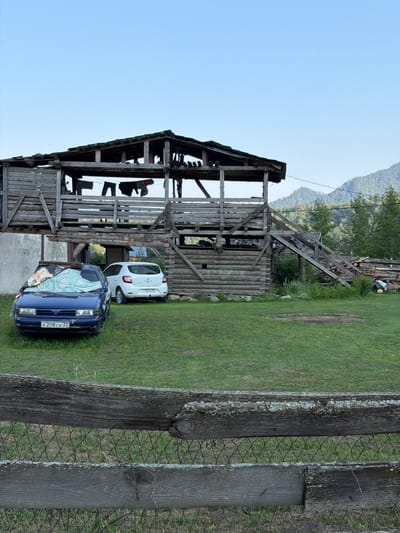 The image depicts a rural scene in the vicinity of Anos, Russia, with a focus on a rustic wooden structure and vehicles. The foreground features a weathered wooden fence with wire mesh, set against a backdrop of green grass. Behind the fence, two cars are parked on the grass. The car on the left is blue and covered with a light-colored tarp, while the car on the right is white.
Dominating the midground and background is a substantial, multi-level wooden structure, appearing to be a barn or storage building. It is constructed from rough-hewn logs and planks, with open sides and a sloped roof, some sections of which appear damaged or incomplete. Laundry is hanging on a line across the upper level of the structure. To the right of the wooden building, a pile of logs is visible, along with some gardening tools.
The environment suggests a mountainous or hilly region, with a forested mountainside visible in the far background under a clear blue sky. The overall impression is one of a functional, perhaps aged, rural homestead in a natural setting. No people are visible in the frame.