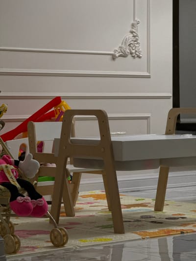 The image depicts an indoor scene in Velikovechnoye, Russia, characterized as a child's play or activity area. The background features a pristine white wall adorned with horizontal classical panel molding and an ornate, scrolled decorative element positioned in the upper right. A dark vertical boundary, possibly a door frame or furniture edge, is visible on the far right.
In the midground, a light-toned wooden children's activity table, topped with a white surface and featuring a central recessed storage tray, is centrally positioned. This table incorporates two open-frame chair-like structures on its opposing sides. Behind the table, partially obscured, is a white child's chair or high chair, and a collection of colorful plastic toys, including prominent red and yellow components, are visible.
The foreground consists of a reflective light-colored tiled floor, partially covered by a multi-colored foam play mat featuring various animal and leaf motifs in pastel shades of yellow, orange, pink, and green. On the left, the lower section of a beige-colored toy stroller with wooden wheels is visible. Inside the stroller, a plush toy identified as Minnie Mouse is seated, wearing a pink dress and black shoes with pink bows, with its white gloved hand making a distinct "thumbs up" gesture. No human individuals are present in the scene.