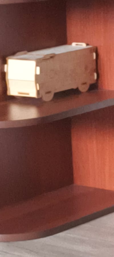 A small, light-colored, toy bus is on a wooden shelf. The bus is made of wood and has simple rectangular windows and circular wheels. It appears to be a miniature replica of a school bus or a similar passenger vehicle. The shelf is made of dark brown wood and is part of a larger shelving unit. The background is a blurred reddish-brown color, suggesting a wall or the back of the shelf. The lighting in the image is warm and somewhat dim, casting a soft glow on the bus and shelf. The scene is devoid of any people or activity. The image lacks specific details that would allow for pinpointing a location within Aktanyshbash, Russia.