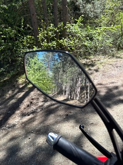 The image shows a motorcycle mirror reflecting a forest path in Boboshevo, Bulgaria. The mirror is mounted on the motorcycle's handlebar, part of which is also visible. The path reflected in the mirror is lined with trees and green vegetation. Outside the mirror, the immediate surroundings include foliage and a dirt or gravel surface with dappled sunlight.