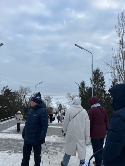 The scene depicts a winter outdoor environment in Primorka, Russia, under an overcast sky. A paved pedestrian pathway, partially covered with snow and ice, descends towards a large body of water in the background, which exhibits visible ice floes or partial freezing. Multiple individuals, dressed in heavy winter clothing, are present on the pathway.

In the mid-foreground, a male subject is visible on the left, wearing a dark blue Columbia puffer jacket, dark pants, dark gloves, and a dark knit beanie with a white pattern; he is turned to his right. Adjacent to him, a female subject is viewed from behind, dressed in a long, off-white, textured coat and light-colored jeans. To her right, another female subject is observed from behind, wearing a maroon puffer jacket and dark pants. A fourth subject, wearing a dark hooded jacket, is partially visible on the far right.

The pathway is flanked by evergreen shrubs and bare deciduous trees and illuminated by several modern streetlights. In the middle distance, additional individuals are visible on the descending path, with at least two subjects carrying inflatable snow tubes or sleds. A metal handrail runs along the left side of the pathway.