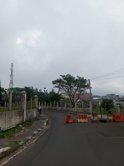 This image depicts a street scene in the city of Houaythat, Laos, under an overcast, uniformly light gray sky.
In the foreground, an asphalt road curves slightly to the right. A faded white line marks the left edge of the road. On the right side of the road, a series of traffic obstructions are present: two orange plastic traffic barriers, an orange traffic cone, two beige plastic traffic barriers, two metal drums (one marked "BR" and the other "08"), and another orange plastic traffic barrier.
Along the left side of the road, a weathered concrete wall topped with a metal fence runs parallel to the curve of the road. Beyond this fence, a grassy area with scattered litter and sparse vegetation is visible. Further left and towards the background, a tall, red and white lattice telecommunications tower stands prominently. Numerous green trees, including some conifer-like varieties, line the property behind the fence. A single individual is observed walking away from the viewer on the paved shoulder adjacent to the concrete wall. This person wears a light-colored head covering, a dark top, blue pants, and carries a bag over their left shoulder.
On the right side of the road, beyond the traffic barriers, a large tree with green foliage stands. Further into the background, utility poles with multiple power lines extend across the upper right portion of the scene. A densely built area comprising various residential and commercial structures with different rooflines is visible, leading towards a large, distant mountain shrouded in mist or cloud cover. One structure in the mid-ground on the right features a distinctive reddish roof.