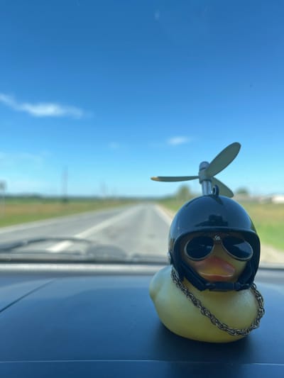 A yellow rubber duck toy adorned with a black helmet, sunglasses, and a gold chain is positioned on a dark dashboard in the foreground. The duck has a propeller attached to its helmet. The background shows a blurry asphalt road stretching into the distance, flanked by green fields and a clear blue sky with scattered clouds. The scene appears to be taken from inside a vehicle, suggesting a road trip. The location in Kukshumy, Russia, is not discernible from the visual information provided in the image.