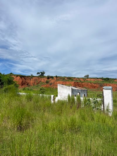 The image depicts an overgrown natural environment containing abandoned structures in Muxara, Mozambique. The foreground is dominated by dense, tall green grass. In the midground, three dilapidated white concrete or masonry structures are partially visible amidst the vegetation. The central structure is a roofless, rectangular building shell with a sloped top and exposed rebar, featuring multiple openings resembling windows or doorways. To its immediate right stands a tall, rectangular white concrete pillar with the words "MUXARA" and "MOÇAMBIQUE" inscribed vertically on its visible side. A partial white wall or foundation is situated to the left of the central structure. The background features a prominent, steeply sloped red earth cliff or embankment, topped with sparse green foliage and a few trees, including a dark green tree near the center. The sky is overcast with a mix of white and light grey clouds. No individuals, animals, or active interactions are present in the scene.