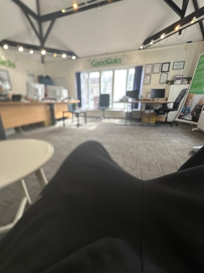 The image depicts the interior of the GoodOaks Home Care office located in Dorchester, United Kingdom, viewed from a seated, first-person perspective. In the immediate foreground, the photographer's legs, clad in dark trousers, are visible, partially obscuring a white, round low table with a metal base to the left.
The midground and background reveal an open-plan office space with a light-colored patterned carpet and white walls. The ceiling features exposed dark wooden beams forming a truss structure, with multiple small, round light fixtures installed. Large windows along the back wall provide significant natural light, looking out onto a street or exterior environment that is mostly blurred.
The name "GoodOaks" is prominently displayed in green lettering on the back wall above the windows. To the left, a light-colored wooden desk is equipped with at least two desktop monitors and associated computer peripherals. Further to the right, another similar desk setup includes a desktop monitor and a black office chair. A series of framed certificates or documents are hung on the wall behind this desk. To the far right, a tall green banner or standee advertising "GoodOaks" services, displaying images of individuals, stands upright. The office appears unoccupied at the time of the photograph.
