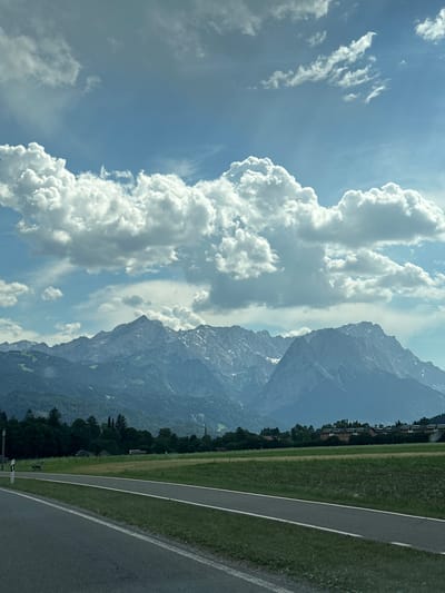An elevated view captures a scenic landscape dominated by a majestic mountain range under a bright, cloudy sky. The mountains, with jagged peaks and patches of snow, form a dramatic backdrop. In the foreground, a paved road leads into the distance, bordered by a grassy field. A dedicated bike path runs parallel to the road. Dense green trees line the foothills of the mountains, and several buildings, including a church spire, are visible in the midground. The overall scene depicts a serene rural setting.