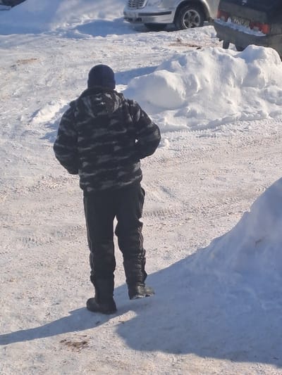 A single individual is depicted walking away from the camera in a snowy, outdoor environment in Bolshaya Elhovka, Russia. The person wears a dark blue beanie, a grey and black camouflage-patterned hooded jacket or sweatshirt, dark pants, and dark boots, with the right foot elevated mid-stride. A distinct shadow of the person is cast on the snow to their left. The ground is covered in snow, showing multiple vehicle tire tracks. In the background, a light-colored SUV, possibly a Mercedes-Benz, is partially visible to the left, and a dark-colored older sedan, possibly a Lada, is partially visible to the right behind a snowdrift. Several snow piles are present throughout the scene, notably a large one on the lower right and another in the mid-ground. The scene is illuminated by bright daylight, casting sharp shadows.