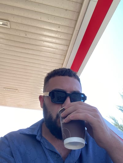 In the city of Korkmaskala, Russia, a man with a beard and sunglasses is seen drinking from a coffee cup. He is wearing a blue button-down shirt with a repeating pattern. The man is positioned under an awning, with a light-colored ceiling above and a bright sky visible. The awning has red and white striped accents.