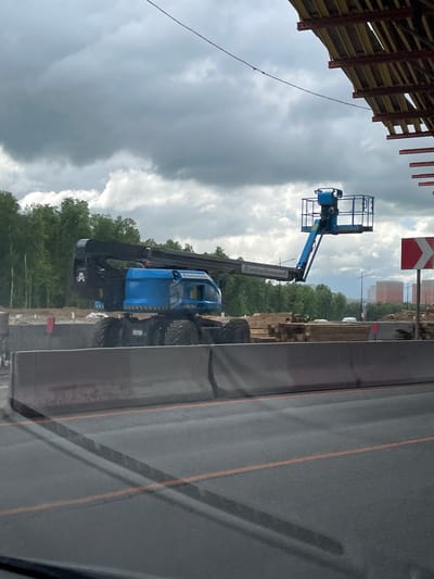 The image shows a Genie S-65 boom lift at a construction site in Novodrozhzhino, Russia. The lift is blue and positioned next to a road. A red and white road sign is visible in the background, along with construction materials and trees. The sky is overcast with dark clouds. A building structure is partially visible on the left side of the image. The photo appears to have been taken from inside a vehicle, with a window frame partially visible on the right.