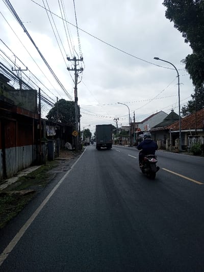 The image displays an overcast daytime street scene in the city of Kamji Na-Yekha, Bhutan. An asphalt road, marked with a solid white line on its left edge and a dashed yellow line in the right lane, recedes into the distance. In the mid-ground, a person wearing a dark blue jacket and a dark-colored helmet operates a dark scooter, moving away from the viewer. Further ahead on the road, a dark green box truck is also proceeding in the same direction.
On the left side of the road, a paved shoulder borders a rusty, corrugated fence or wall, above which a concrete structure is visible. A utility pole heavily laden with a dense network of overhead electrical wires stands prominently on the left. Further down, a yellow triangular road sign with a curved arrow is partially discernible. Buildings, featuring various roof styles including terracotta tiles, line both sides of the street, suggesting a mix of residential and commercial properties. On the right side, another utility pole with a curved street light arm is present. Green foliage from trees partially obscures some buildings and the sky on the far right. The sky is uniformly overcast.