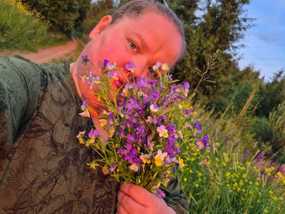 A person is holding a bouquet of small purple and yellow wildflowers in the foreground of the image. The person's face is partially visible, tilted and close to the flowers, with their skin flushed red from the evening sun. They are wearing a green, patterned shirt. The background shows a grassy field with more wildflowers and trees, suggesting an outdoor, rural setting, possibly near Doinaya, Russia, given the context. The sky is blue with some clouds, and power lines are visible in the distance. The image is a selfie taken at golden hour.