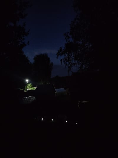 The image depicts a nighttime scene with minimal illumination, suggesting a rural or outdoor setting, possibly in Beryozovka, Russia. The sky is a deep, dark blue, hinting at twilight or a moonless night. Silhouetted trees frame the top and sides of the image, their branches creating an intricate, dark pattern against the sky.

In the lower right portion of the frame, a series of small, bright lights are visible, arranged in a vertical line. These appear to be outdoor string lights or a similar decorative lighting setup. Further down and to the left, a solitary, brighter light source illuminates a patch of greenery, possibly a bush or small tree. The overall impression is one of a quiet, somewhat serene night, perhaps with a gathering or an event taking place outdoors, marked by the presence of the lights. No people are clearly identifiable, though shapes suggest a possible presence in the darker areas. The low light conditions obscure most details, creating a mood of mystery and nighttime ambiance. There is no visible text in the image.