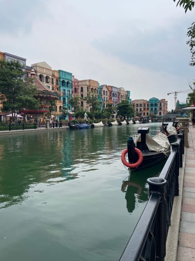 The image depicts a canal lined with colorful buildings in the city of Nghĩa Trụ Commune, Vietnam. The architecture appears to be inspired by European styles, with pastel-colored facades and ornate details. Several gondolas, covered with white tarps, are moored along the canal, suggesting a tourist attraction or a themed area. One gondola in the foreground is uncovered, showing a red life preserver. People are visible on the walkway beside the canal, some standing near a carousel. The water in the canal is a deep green, and its surface reflects the buildings and sky. A black metal railing runs along the edge of the walkway. In the background, a construction crane is visible, indicating ongoing development. The sky is overcast, with a muted, diffused light. Trees and greenery are interspersed between the buildings.
