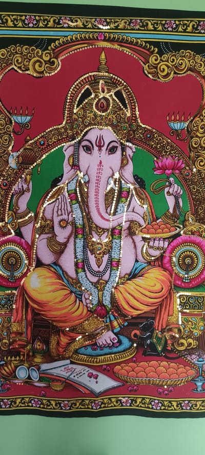 This image is a detailed, colorful painting of the Hindu deity Ganesha. Ganesha is depicted with his characteristic elephant head and is seated in a cross-legged position. He has multiple arms, some of which are holding objects such as a lotus flower and a dish of sweets. The painting is richly adorned with gold sequins, which catch the light and add a shimmering effect. Ganesha is wearing ornate jewelry and vibrant clothing. The background of the painting features intricate decorative elements and is predominantly red and green. At the bottom of the painting, there are offerings including coconuts and a plate of sweets. The style of the painting is traditional Indian art. There is no discernible information within the image that indicates its location in the city of Смородинка Дп, Russia.