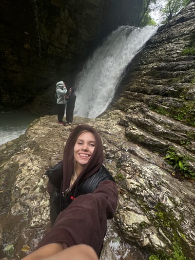 A woman in a brown hooded sweatshirt and black jacket is taking a selfie at the "Bagrat's Waterfall" location in Tsabal, Abkhazia. Two other people are visible near the base of the waterfall in the background. The environment consists of rocky cliffs, vegetation, and flowing water.