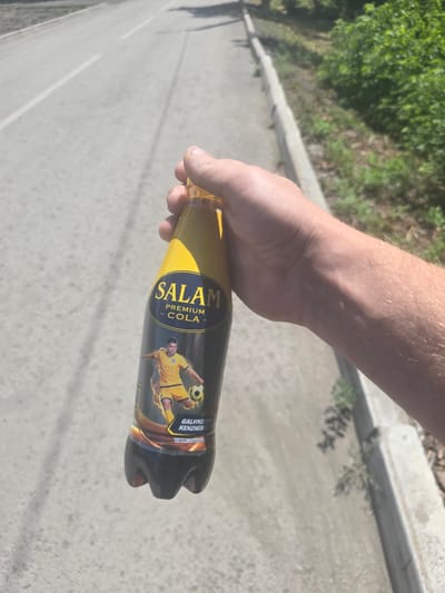 A hand holds a bottle of "SALAM PREMIUM COLA" with a soccer player depicted on the label, in front of a paved road with a curb and vegetation. The bottle is dark with a yellow label. The road appears to be in a sunlit outdoor environment, possibly a street or walkway in the city of Saran, Kazakhstan, given the context. The image is taken from a slightly elevated perspective, looking down at the bottle and the road.