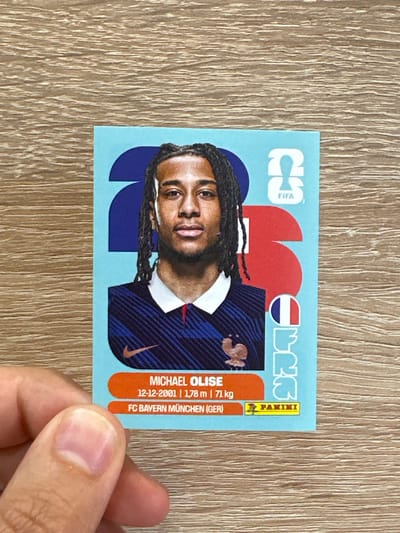 A sticker featuring a portrait of a man with dark, braided hair and a blue and white striped shirt is held by a person's hand. The sticker has a light blue background with large, abstract red and blue shapes behind the man. Text below the portrait reads "MICHAEL OLISE 12-12-2001 | 1,78 m | 71 kg FC BAYERN MÜNCHEN (GER)" and "PANINI". A small image of a rooster is also visible on the sticker. The sticker is placed on a wooden surface. There is no discernible location in Prisovo, Bulgaria.
