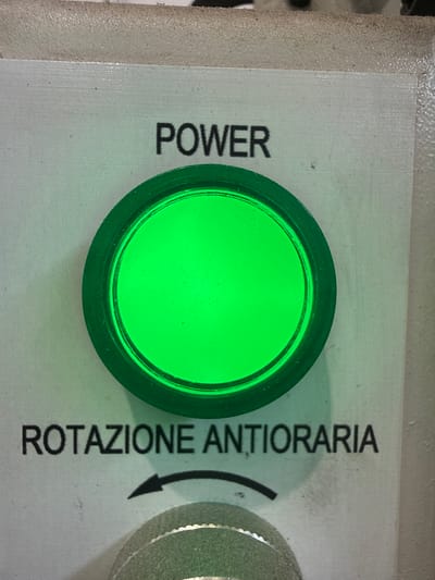 This is a close-up, eye-level, overhead shot of a control panel that reads "POWER" at the top and "ROTAZIONE ANTIORARIA" below. A bright green indicator light is illuminated in the center of the panel, positioned between these two text labels. Below the "ROTAZIONE ANTIORARIA" text, there is a black arrow pointing counter-clockwise. The panel appears to be made of a light gray metal with some surface texture and a few small specks of dirt or debris. The lower part of the image shows a portion of a round, silver-colored knob with a textured edge. The context of this image is not related to the city of Fontevivo, Italy.