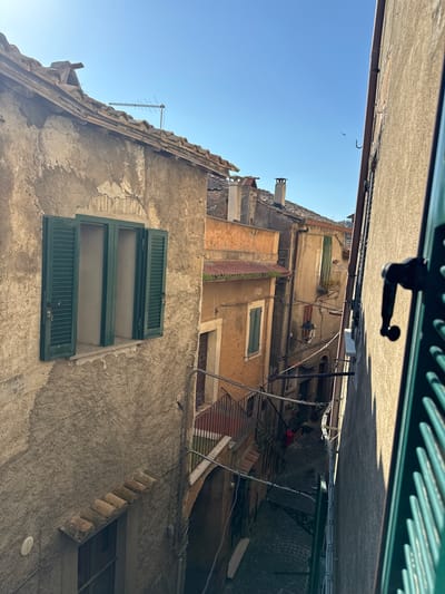 The image shows a narrow street in Castelnuovo di Porto, Italy, looking upward between buildings. The buildings are old, with weathered facades, and feature exposed brickwork, aged plaster, and green shutters on the windows. An antenna is visible on the roof of one building. Overhead, a small gap reveals a clear blue sky. At the top of the frame, a green metal grate and a window handle are visible, suggesting the photo was taken from inside a building looking out. The street below appears to be paved with cobblestones, and there are various wires and pipes running along the buildings' exteriors.