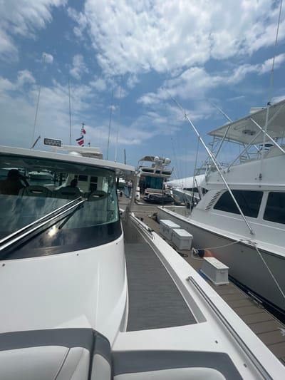 The image shows a white boat docked at a marina in Playa Herradura, Costa Rica, a popular tourist destination known for its beaches and fishing. The boat is positioned at the marina, which is characterized by a wooden dock. The boat is white with a gray stripe along the side, and it is clean and well-maintained. The boat's windshield is dirty, but it is not clear if it is due to rain or dust. There are several other boats in the background, but none are visible in detail. The image focuses on the boat's front end, highlighting its features, such as the windshield, the side rails, and the deck. The sky is clear and blue with scattered white clouds. The Costa Rican flag is visible on the boat's mast.