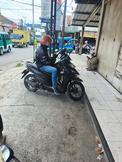 A person on a black motorcycle wears an orange helmet and blue jeans. The motorcycle is parked on the sidewalk, next to a building with closed, peeling wooden doors. A person wearing a helmet sits on the ground near the building, appearing to be using a phone. Vehicles pass on the street behind the motorcycle. Trash is on the sidewalk, including small plastic bags.