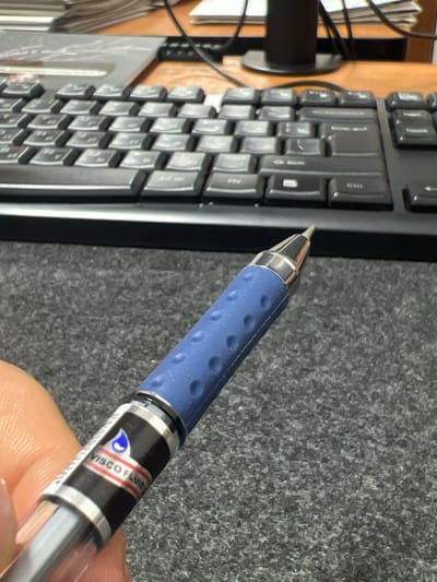 A close-up, angled shot shows a blue and silver pen in the foreground, held by an out-of-focus left hand. The pen has a textured blue grip and a silver tip. A black and white label on the pen barrel reads "VISCO FLUID" with a blue droplet logo. In the background, a black keyboard is visible, with some keys appearing to have Cyrillic characters. A stack of papers sits on the desk behind the keyboard. The environment appears to be an office or study setting, with a wooden desk surface and a speckled grey carpeted surface. The image does not provide any specific geographical information that would allow for its location to be identified as Kadamjay, Kyrgyzstan.