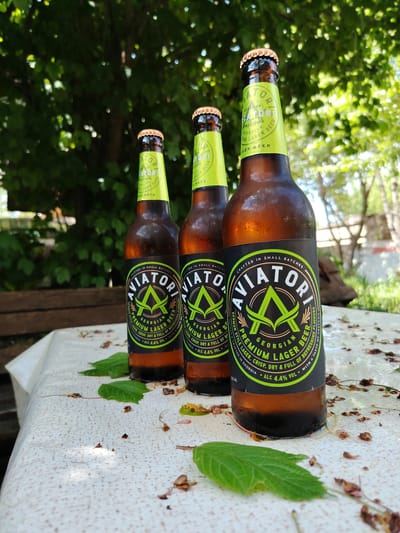 Three bottles of Aviatori Georgian Premium Lager Beer are arranged on a table outdoors. The table is covered with a light-colored, patterned cloth and is littered with fallen leaves and small twigs. The bottles are amber glass and have prominent black and neon green labels. The background consists of blurred green foliage, suggesting a natural, possibly wooded, setting. The image is shot from a low angle, with the bottles in sharp focus and the background softened. The lighting suggests it is daytime, with dappled sunlight filtering through the leaves. There are no people visible in the image. The setting is consistent with an outdoor dining or picnic area in Bakuriani, Georgia.
