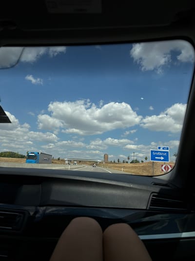 The image is taken from inside a vehicle, looking out the windshield. The sky is blue with fluffy white clouds. A highway is visible in the distance, with cars and trucks driving on it. There is a blue and white road sign that reads "41" and "Großkrut" with an arrow pointing to the right. There is also a red and white directional sign. To the left, a blue truck is visible. The foreground shows the car's dashboard and the legs of a person. The scene appears to be on a highway in Poysdorf, Austria.