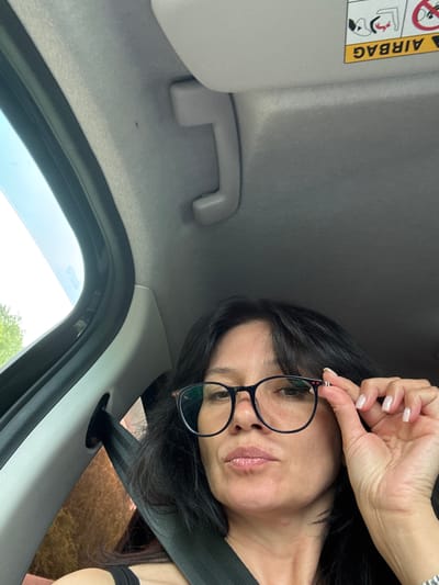 The image shows a woman in a car, adjusting her glasses. She has dark hair and is wearing a black tank top and glasses. The car interior is visible, including the headliner, a sun visor with an airbag warning label, and a seatbelt. The view outside the car window shows trees and sky. The provided location, Kazaevo, Russia, is not discernible from the visual information in the image.