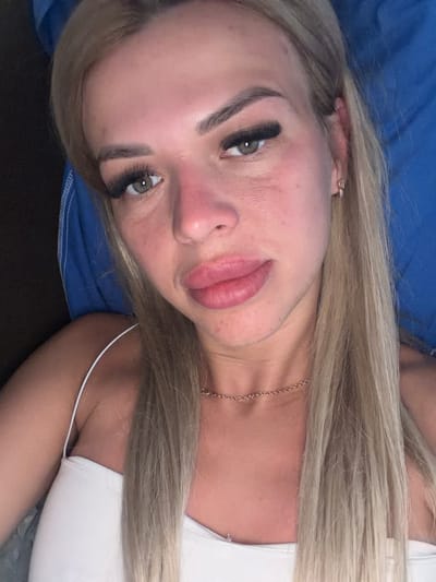 A close-up selfie of a fair-skinned blonde woman is shown, taken from a high angle. She is wearing a white top and a thin gold chain necklace. Her hair is long and blonde, falling to her shoulders and over her chest. She has light green eyes, heavily made-up with thick false eyelashes. Her lips appear plump, and the skin on her face shows some redness and texture. She is lying down, with a blue pillow visible behind her head and a dark surface to her left. There is no indication of her location within the city of Kyiv, Ukraine.