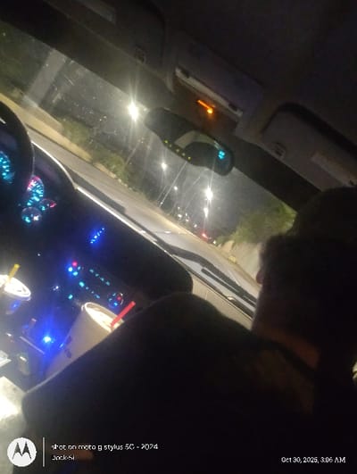 The image depicts the interior of a vehicle at night, viewed from a low-angle perspective looking forward through the windshield. The timestamp indicates "Oct 30, 2025, 3:06 AM."

**People:** The head and right shoulder of an individual are visible in the foreground on the right side, suggesting a passenger. A driver is implied by the presence of a steering wheel, partially visible on the left side of the frame.

**Subjects/Objects (Vehicle Interior):**
- A dashboard is prominent on the left, displaying multiple blue-illuminated gauges, including a speedometer and tachometer with bright blue needles.
- Two cups are situated in the center console cupholders: one is a white Sonic Drive-In cup with a red lid and red straw, and the other is a lighter-colored cup with a yellow lid and yellow straw, partially obscured.
- A rearview mirror is visible in the upper center, featuring a digital display (possibly "N 32" for north and temperature) and several small green indicator lights on its lower edge.
- An overhead console contains an illuminated orange indicator light.

**Subjects/Objects (Exterior Environment):**
- Through the windshield, a multi-lane road is visible, brightly illuminated by numerous streetlights that cause significant lens flare.
- Faint red tail lights of other vehicles are discernible further down the road.
- Dark vegetation and what appears to be a concrete barrier or retaining wall line the left side of the road.

**Actions/Interactions:** The vehicle appears to be in motion or stopped on a road at night.

**Location:** The scene is set in an urban or suburban area within Tulsa, United States. However, the precise street or highway location within the city cannot be determined from the visual details provided in the image.