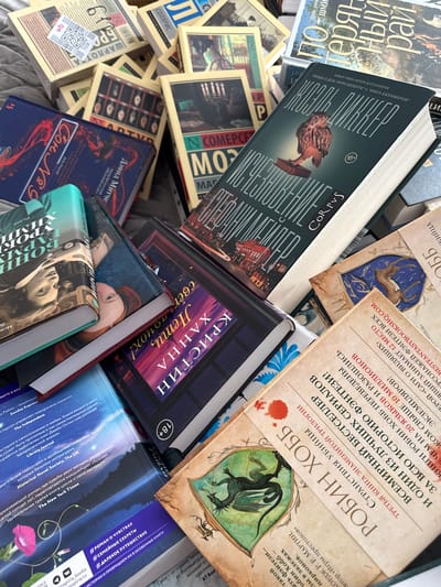 The image shows a pile of Russian books. The books vary in size and cover design. Some titles visible are "Corpvs," "Son No. 9," "Kristin Hanna: Leti, svetlo," and "Robin Hobb." There is a book with a picture of an owl on its cover. The image is taken from above. The book titles and snippets of text indicate a focus on fantasy, historical, and contemporary fiction. The variety in cover design suggests a mixture of genres and authors.