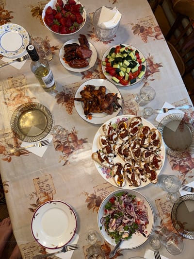 The image depicts a table set for a meal, likely outdoors given the natural light. The table is covered with a tablecloth featuring a vintage botanical print with script text. On the table, various dishes are arranged: a bowl overflowing with fresh strawberries, a plate of grilled meats including sausages and what appear to be chicken wings, a salad composed of chopped tomatoes, cucumbers, and red onions, and a platter of bruschetta topped with a creamy spread and a dark drizzle. A bottle of white wine and several glasses are also present, suggesting a gathering for a meal. The overall impression is one of a relaxed, celebratory occasion. There are no visible people in the image, but the setting suggests a meal is about to be or has been consumed. The environment appears to be a domestic setting, possibly a patio or garden area, given the ambient lighting and the presence of a wooden chair in the background. No specific landmarks or distinguishing features of Усть-Мана, Russia, are visible in the image.