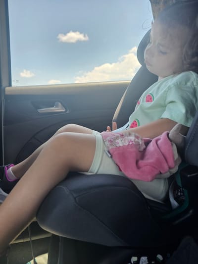 A young child is seated in a car seat, facing away from the camera and towards the right side of the frame. The child appears to be asleep or resting, with their eyes closed and head tilted slightly downwards. They are wearing a light green t-shirt with a watermelon pattern and light-colored shorts. The child is holding a pink blanket or stuffed toy.
The car's interior is visible, including a dark gray car seat, door panel with a silver handle, and a portion of the car window showing a bright blue sky with scattered white clouds. The ambient lighting suggests it is daytime.
There is no specific indication within the image that places the scene in Staro Selo, Bulgaria. The provided image is a generic interior car scene.