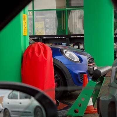 The image captures a scene from inside a vehicle, looking out at a service station or car wash bay. A large, red, crinkled object, possibly a bag or car cover, occupies a significant portion of the foreground. Beyond this, a prominent green vertical structure frames the left side of the view.
A blue car, identifiable as a Mini Cooper by its distinctive headlight and grille design, is parked in the middle ground. Its front-left wheel is visible, revealing a red brake caliper with "WORKS" branding. Two orange traffic cones are positioned around the car, suggesting a designated area or service bay. To the right, part of a green metal structure with circular cutouts is visible, with another orange cone behind it. Behind the blue car, shelves stocked with various bottles, likely automotive fluids, are arranged.
The scene includes a large window reflecting the interior of a building. Prominently visible in the reflection is a yellow sign with text in both Russian and English: "СПАСИБО, ЧТО ОПЛАТИЛИ ПРОЕЗД" and "THANK YOU FOR TOLL PAYMENT," accompanied by the "BelToll" logo. Other partial Russian texts from the OCR include "ВНИМЕ" (Attention), "нефть" (oil), "вам", "прав", "Вните", and "TK". The overall lighting suggests it is daytime. The setting appears to be a commercial service area for vehicles, situated in Дорскі сельскі Савет, Belarus.