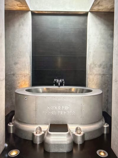 This media file shows a large, circular, metal vault door, set within a concrete room. The vault door appears to be made of brushed aluminum or stainless steel, with visible rivets and a textured surface. It has a thick, imposing frame and a central locking mechanism with multiple bolts. The room itself has textured concrete walls and a dark, tiled floor, giving it a modern, industrial aesthetic.

To the right of the vault door, there is a small, recessed circular light fixture emitting a warm, yellow glow. There are also some visible markings on the vault door itself. These markings appear to be embossed or etched into the metal and are somewhat difficult to decipher clearly. They seem to be a combination of letters and symbols. The image is taken from a high angle, looking down at the vault door and the surrounding room.

The lighting in the scene is a mix of artificial light from the fixture and ambient light that casts soft shadows, creating a somewhat dramatic and imposing atmosphere. There are no people visible in the image, and the scene appears to be static, with no discernible activity taking place. The overall impression is one of security and industrial design.

The location context provided is Borowa, Poland, which could suggest this is a vault in a bank, a secure facility, or perhaps an art installation. The time of day and weather conditions are not discernible from the image.