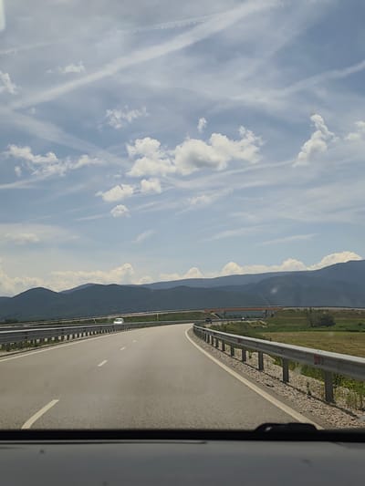 The image presents a view from inside a vehicle traveling on a multi-lane highway near Skravena, Bulgaria, captured from what appears to be the driver's or front passenger's perspective. The asphalt road curves gently to the right, marked with white dashed and solid lines. A metal guardrail runs along the right edge of the highway, beyond which lies a grassy roadside and a wider green landscape.

In the mid-distance, an orange-brown bridge or overpass spans across the scene. A white car is visible on the highway further ahead, and another dark vehicle can be seen underneath the distant overpass. The background is dominated by a range of dark, rolling mountains or hills, indicating a mountainous or hilly terrain.

The sky is bright blue, adorned with scattered white cumulus clouds and several distinct, linear contrails (likely from aircraft) crisscrossing the upper sky. The weather appears clear and sunny, suggesting a pleasant daytime journey. No people are explicitly visible, nor is any text present in the scene. The overall impression is one of open road travel through a scenic, mountainous region.