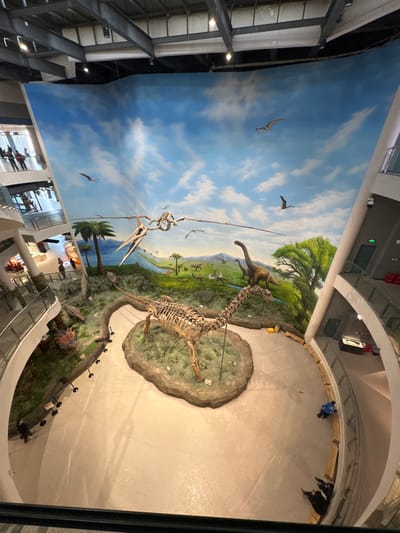 The image shows a museum exhibit from an elevated perspective. A large diorama is visible, featuring a painted backdrop with dinosaurs and pterodactyls. Below, on a raised platform, stands a dinosaur skeleton and a model of a dinosaur. People are visible on the floor above and in the levels below the diorama. The building's architecture includes exposed metal supports and glass railings.