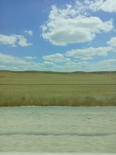 This is a photograph taken from a vehicle, likely in motion, showing a vast, flat agricultural landscape under a bright blue sky dotted with fluffy white clouds. The foreground features a blurred expanse of what appears to be a road or a light-colored ground surface. Beyond that, a horizontal band of dried, yellowish-brown vegetation stretches across the frame, possibly a harvested field or grassland. In the distance, rolling hills with a green hue form the horizon line, suggesting gentle undulations in the terrain. Thin power lines are visible stretching across the upper portion of the sky, indicating the presence of infrastructure. The overall impression is of a rural, open environment. There are no people or specific landmarks to identify the exact location within Balâ, Turkey.