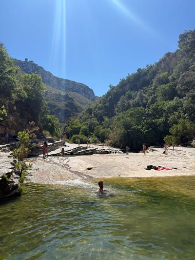 The image depicts a natural swimming area with a shallow river flowing over a rocky bed. Several people are visible, some swimming in the river and others relaxing on the sun-drenched rocky banks. The surrounding environment is lush and green, with trees and foliage covering the steep hillsides that form a gorge. The sky is clear and blue, with the sun casting bright rays, creating a vibrant and inviting atmosphere. Based on the provided context that this location is in the city of Noto, Italy, this scene is likely the Calamosche beach or a similar natural pool within the Vendicari Nature Reserve, known for its pristine beauty and crystal-clear waters.
