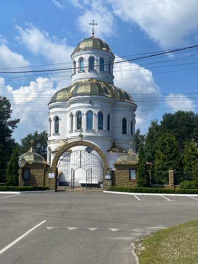 A white, domed Orthodox church with golden accents stands behind an ornate wrought-iron gate. The church has multiple levels and arched windows, topped with a cross. The gate is set within a stone archway, flanked by two smaller golden domes on stone structures. Trees surround the church, and a paved area with parking lines is visible in the foreground. The sky is blue with scattered clouds. This is the Church of the Transfiguration in Liutizh, Ukraine.