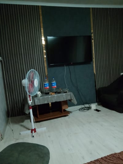 The image depicts an indoor living space, possibly a lounge or bedroom, with a dark, textured wall featuring vertical stripes and a narrow gold accent. A large flat-screen television is mounted on this wall. To the left, a white oscillating floor fan with red accents stands on a light-colored floor. In front of the fan is a small table covered with a gray cloth, on which sit two bottles of cola, two glasses with dark liquid, and a white mug. A dark sofa is visible to the right of the television. The floor is a light wood laminate. Electrical cords and a power strip are present on the floor near the sofa. The scene appears to be set in a residential dwelling within the city of Кугейский, Russia.