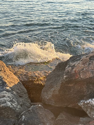 A close-up, eye-level shot captures a wave crashing against rocks at the shoreline in Alimos, Greece. The water is a deep blue-green with visible ripples and white foam from the breaking wave. The rocks are varied in size and color, with shades of gray, brown, and hints of red. Some rocks are wet and appear to have algae growth. The sunlight casts shadows and highlights on the textured surfaces of the rocks, indicating it is likely late afternoon or early morning. The scene is devoid of human presence.