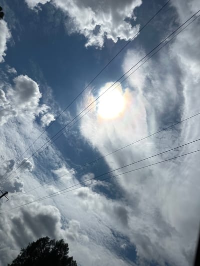 The image depicts a view of the sky with clouds and power lines. The sun is visible, partially obscured by clouds, creating a halo effect with iridescent colors. The clouds are varied in texture and density, with some appearing bright white and others darker and more shadowed. Four parallel power lines run diagonally across the frame from the upper left to the lower right. At the bottom left, the silhouette of a tree is visible, along with a portion of a utility pole. The scene appears to be an outdoor shot taken on a sunny day with scattered cloud cover. There are no people or animals visible in the image. The location is specified as Новая Казмаска, Russia.